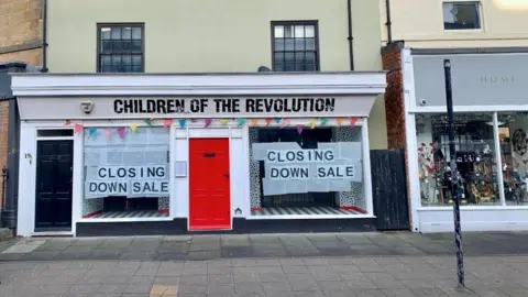 View of a double fronted shop, Children of the Revolution. The windows on either side of the red door have signs reading "closing down sale".