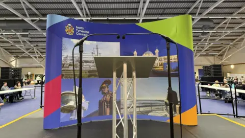 The small black stage has two microphones and a podium on the front. There is a large North Tyneside Council banner behind with various pictures of the area, including the white domes of Spanish City.