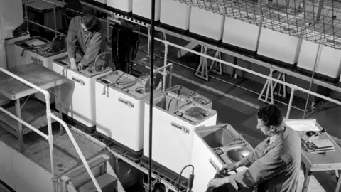 Getty Images Two men in boiler suits working on white washing machines which are lined up in rows.