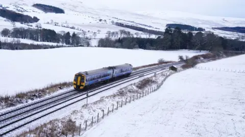 PA Media A train travelling through countryside, with snow covering the hills and grounds around it