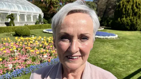 Siobhan smiling at the camera. She is wearing a light pink jumper and is standing outside on a sunny day. She has short grey hair. Behind her there are gardens with colourful flowers.