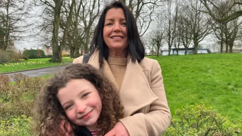 Louise James stands behind her daughter Rioghnach. Louise has long dark hair and is wearing a tan woollen coat over a tan woollen sweater. She has her arms on Rioghnach who is smiling and has curly brown hair. They are standing in a park, trees and a rolling stretch of grass can be seen behind them.