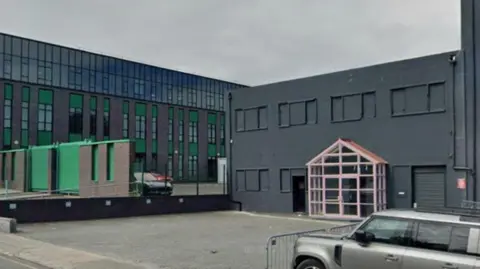 A street view of the site which is a grey building with boarded up windows. The entrance has a glass front. A big grey, green and glass building can be seen to the left.