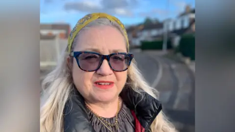 Sarah Massey has long blonde hair and a yellow hairband. She is standing on the pavement with a road behind her.