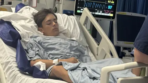 Finley lies in a hospital bed, his eyes closed, a screen displaying his vital signs behind him.