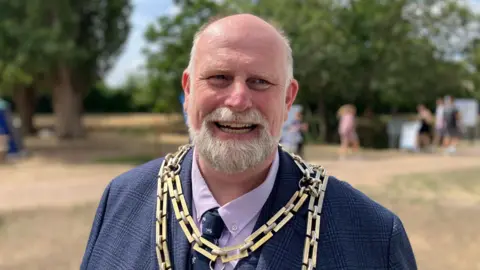 Sam Read/BBC Tom Wootton wears a ceremonial chain of office and dark jacket and stands outdoors in a park-like setting. Trees and a small number of people are visible in the blurred background beneath a bright sky.