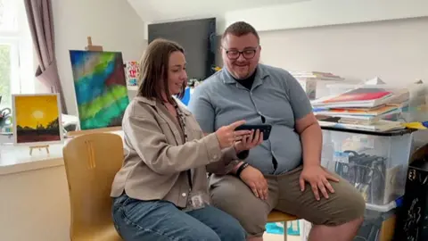 A man in blue top and khaki shirts and glasses looking at mobile phone footage held by a woman in blue jeans and beige top. She has long brown hair