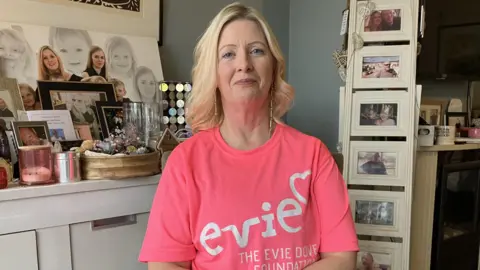 A woman with blonde hair sat in a living room in a house surrounded by photographs of her family. She is wearing a bright pink T'shirt and her hands are on her lap. 