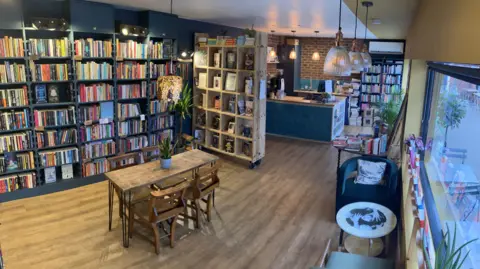 The Heath Bookshop The inside of a book shop with shelves along the backwall filled with books. There is a large shopfront window on the right with seating underneath it. In the middle of the room there is a wooden table and chairs. At the back of the room are more shelves and a coffee bar