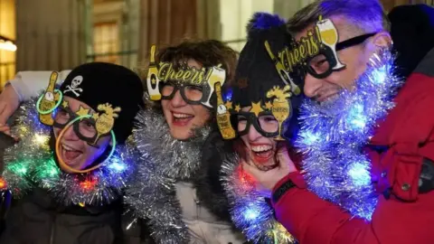 PA Four revellers wearing funny glasses and tinsel. They are all laughing. Two are wearing dark woolly hats.