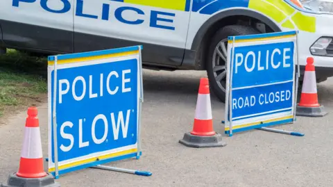 Two bright blue police signs are at the side of the road. One reads "police, slow" and the second says "police, road closed". There are three orange cones also at the side of the road, and a police van is visible in the background. 