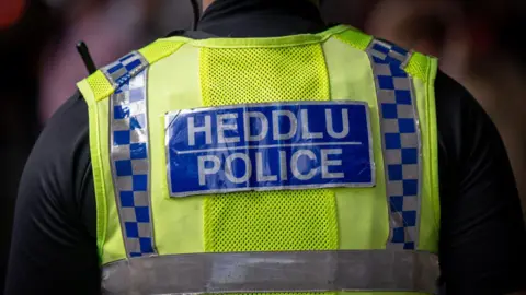 The back of a police officer in Wales. He is wearing a neon yellow vest over a black top. The vest has a blue box with grey writing with 'Heddlu Police'