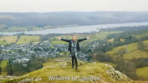 Coniston Parish Council A screenshot of the promotional video featuring a woman standing on the top of a fell with Coniston in the background. The subtitles read "Welcome to Coniston".