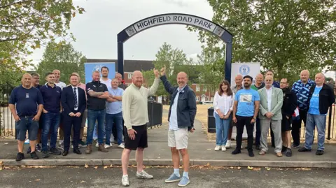 Groups of people including Jim Brown and Andy Whing who are pointing up towards the Highfield Road Park sign at the park.