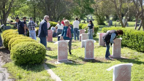Commemorative Air Force Memorial stones on grass with tracing paper over them. The tracing paper has been coloured in with crayon to show the design and writing on the stone. There are several people walking around the stones looking at them.