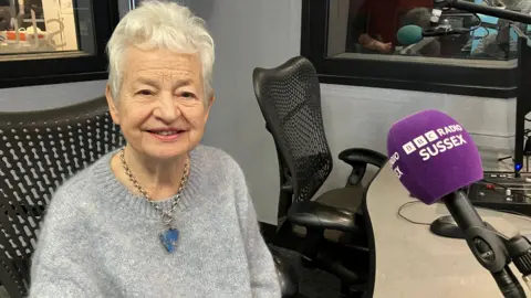 Jacqueline Wilson sat in a radio studio for BBC Radio Sussex. She is wearing a light blue jumper and a big blue necklace, she has grey hair. 