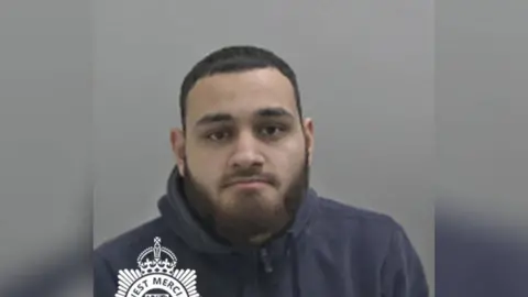 A mugshot of a man with short black hair and a dark beard. He is wearing a black hoodie and looking into the camera.