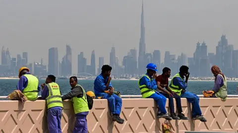 Getty Images Workers wearing blue uniform and neon green jackets resting on the beardwalk with the Dubai skyline in the background during daytime