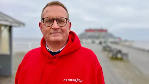 Martin Giles/BBC A man wearing a red hoodie with Cromer Pier written across the left side of his chest looks at the camera. He has short brown hair and is wearing brown-framed glasses. 