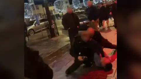 A screenshot from a video shows three individuals on the ground - two police officers who appear to be restraining a member of the public - while several people look on. The faces are blurred. The shot is taken on a street at nighttime. 