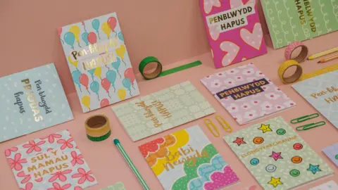 ওয়েলশ ভাষার জন্মদিনের কার্ডগুলি গোলাপী টেবিলে বসে আছে। তারা পেনউইড হাপুসের মতো কথা বলে। ওয়েলশ জন্মদিনের শুভেচ্ছা। কার্ডগুলির মধ্যে রঙিন কাগজের ক্লিপ, মোড়ানো ফিতা এবং পেন্সিল রয়েছে।