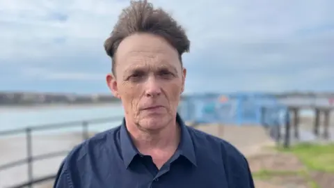 Paul Jenkins is clean shaven with medium-short brown hair blowing back off his face in the wind and is wearing a navy shirt. Behind him, but out of focus, are some railings and, beyond those, a beach and the sea. A small patch of grass is just visible, though blurred, immediately behind him and to the right.
