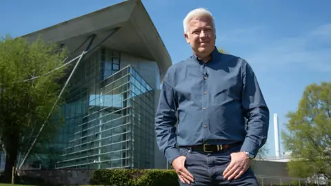 BBC news A man stands in front of a glass building (Met Office in Exeter) with his hands in his pockets. He is wearing a navy shirt and jeans