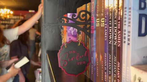 A bookshelf with a small black sign stuck among the books, saying The Sinful Spine in cursive red script. In the background, people can be seen reaching for the books, but they are out of focus.