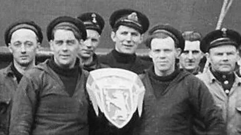 A black and white picture of a group of about 18 men and two women. They stand shoulder to shoulder in a line, with some of the men on one knee in front of the group. Most wear navy caps.