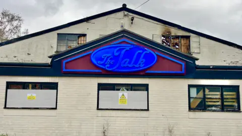 Martin Giles/BBC Part of The Talk, a former nightclub on Oak Street in Norwich. The building is a brick building which has been painted white. There is a blue sign with cursive writing which says, The Talk, on it. There is smoke damage from a fire on the windows of the roof of the building.