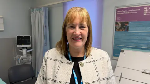 A woman wearing a light-coloured, checked jacket and an NHS lanyard stands in a hospital room. Medical equipment, a curtained area and information boards are visible in the background.