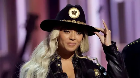 Getty Images Beyoncé accepting an award on stage, wearing a black cowboy hat, using her left hand to hold the top of the hat. Her black outfit also has golden buttons.