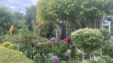 Katie Jaffar A beautiful front garden can be seen. It is filled with trees, flowers, plants and sculptures. Some purple alliums can be seen in the foreground. The sky behind is cloudy A hand-made ladybird can be seen on the trunk of one of the trees.