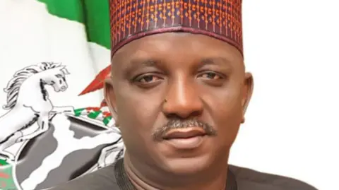 Saleh Mamman poses looking directly into the camera. He wears a grey top, a multi-coloured cap, and is pictured in front of a flag.