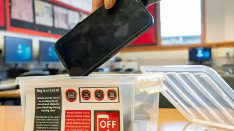 PA A picture of a student's hand placing a mobile phone in a plastic box in a school