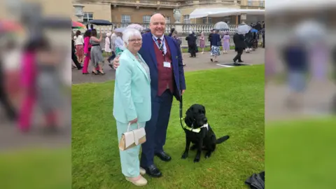 John Hardy with his arm around his wife's shoulders. They are both in suits standing in the garden of Buckingham Palace. John is holding Sidney, a black Labrador, on a lead.