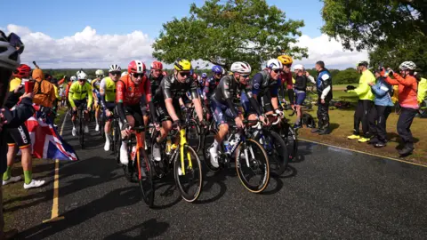 PA Media A crowd of racing cyclists wearing helmets and T-shirts of different colours. They are passing a small number of spectators who are standing on the grass next to the road. There are trees and clouds in the background.