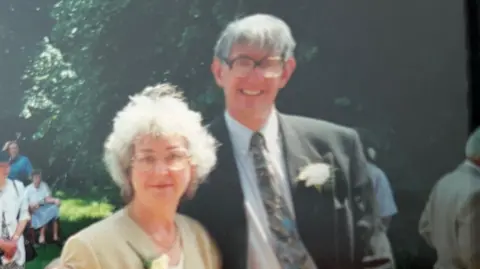 An old colour photo of Jean Turnbull and Raymond Turnbull, taken outside at a wedding or similar event. They both have large glasses and flowers in their button holes. Jean has white-grey hair to just above the shoulder and is wearing a yellowish tan light jacket. Raymond has grey hair brushed forward and wears a dark grey suit jacket over a very pale blue shirt and patterned tie. It is a sunny day and a large tree thick with leaves forms the background. A few other guests can be seen milling around.