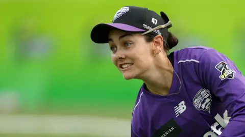 Heather Graham in action wearing a purple long sleeve kit and a black cap with a purple peak. Her sunglasses are on the back of her cap and she has a long gold chain around her neck.