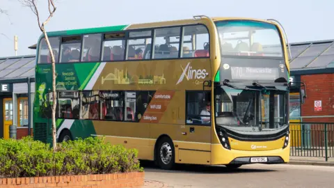 A yellow and green double decker X70 bus to Newcastle leaving a bus station