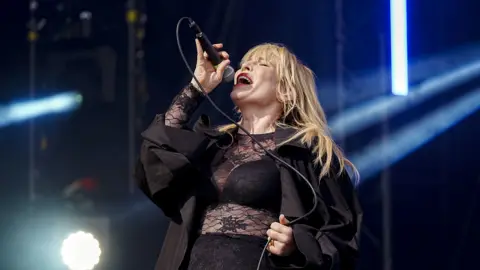 Natasha Bedingfield, wearing a black lacy outfit, closes her eyes and sings into a microphone. She has long blonde hair draped on her shoulders. 
