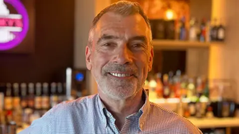 Brett Martin, a man with short grey hair, who wears a chequered navy and white shirt. He stands at a bar and smiles.