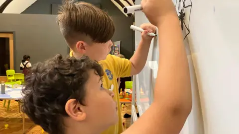 Two students in the class are drawing on the blackboard.  The boy closest to the camera has curly brown hair and is drawing a spider using a black pen.  His classmate had his hair partially shaved and was drawing with green ink.