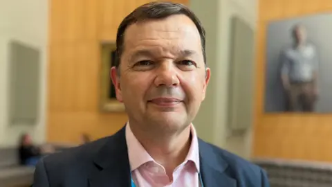 Prof McIntosh looks directly at the camera. He has short brown hair and wears a pink open-necked shirt under a blue suit jacket