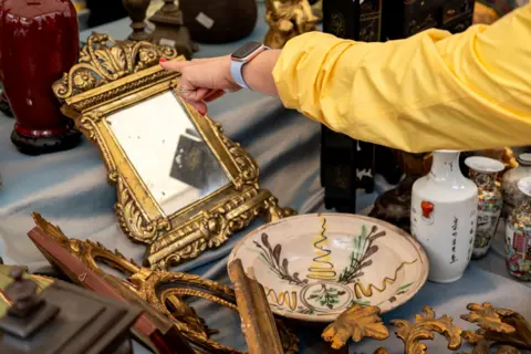 Getty Images A shopper in a yellow jacket points at an ornate gilt mirror among vintage ceramics and decorative frames on a flea market stall.