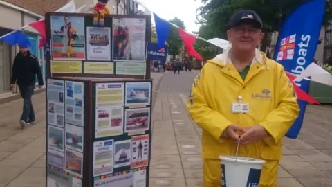 RNLI Clive Buckley wears a yellow RNLI jacket and holds a bucket to collect money from passers-by. There is RNLI bunting and a flag behind him.