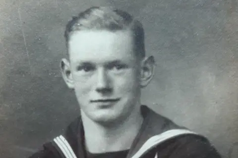 The Redston Family Bill Redston in a black and white image dressed in a Royal Navy uniform. He has short, dark hair with a side parting and is clean shaven.