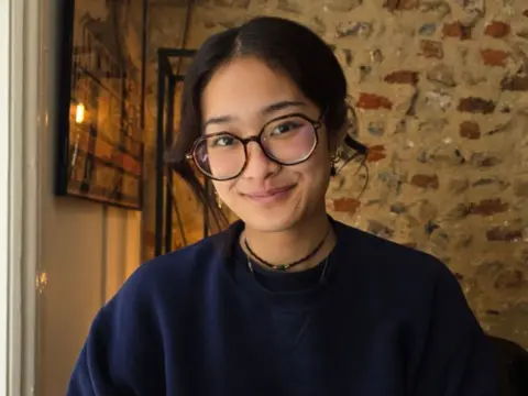 Pia Subong A woman in a blue jumper and dark glasses smiles in front of a brick wall