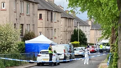 BBC A cordoned off area of a street in South Queensferry. There are people wearing white hazmat suits and uniformed police officers. A blue forensic tent is in the front garden of one of the properties and cars line the street.