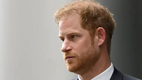 Prince Harry in a suit with a black blazer and tie. He has ginger hair and beard, and also blue eyes. He is looking off into the distance.